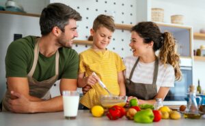A family of three, including a man, a woman, and a child, are gathered in a bright kitchen, preparing food together. The child is stirring ingredients in a bowl while the adults watch and smile. Fresh vegetables and a glass of milk are on the counter.